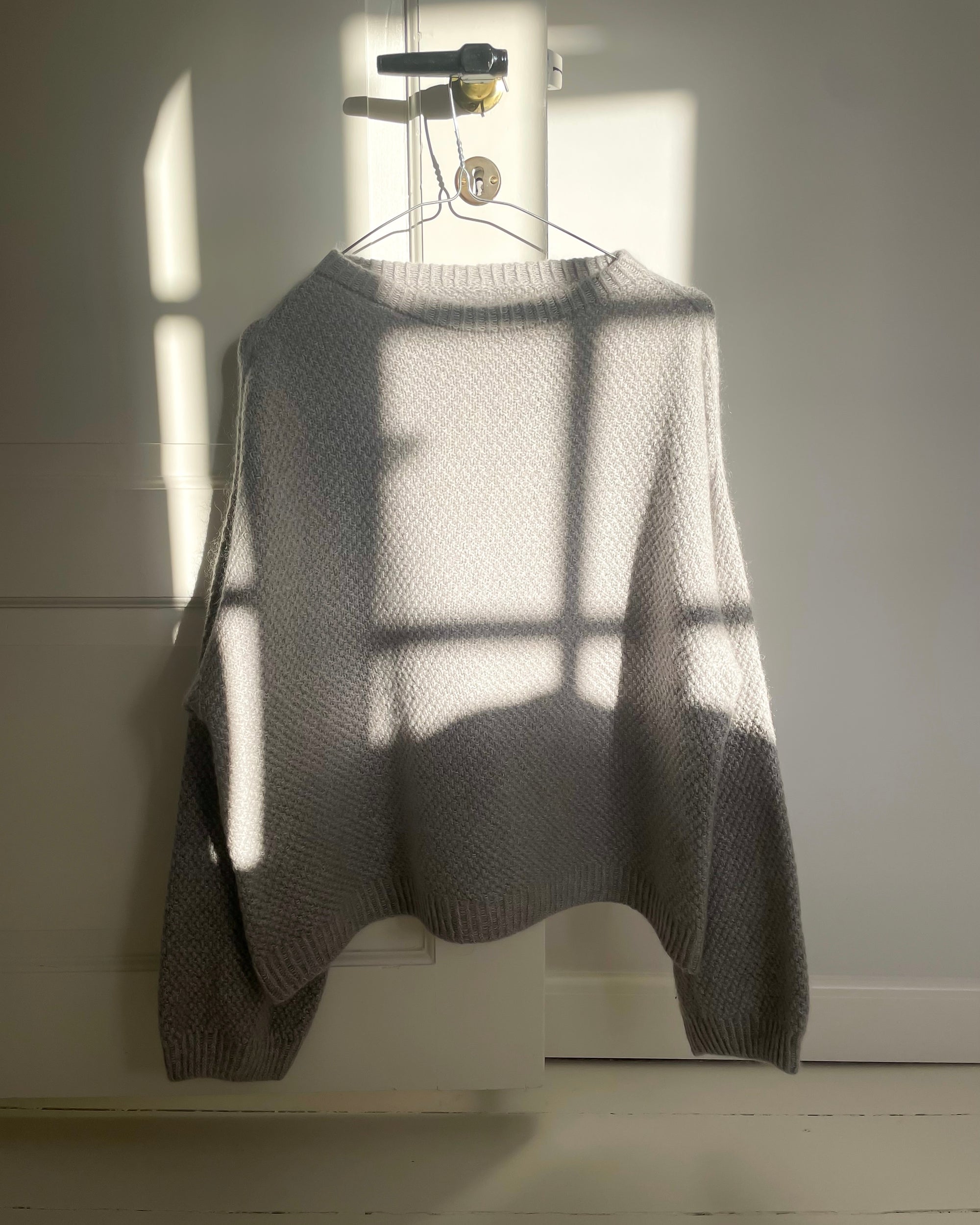 Photo of a light grey version of the Bondi Sweater hanging on a door handle bathed in sunlight. Bondi Sweater is a hand knit sweater by Berthyknit worked in double moss stitch. 